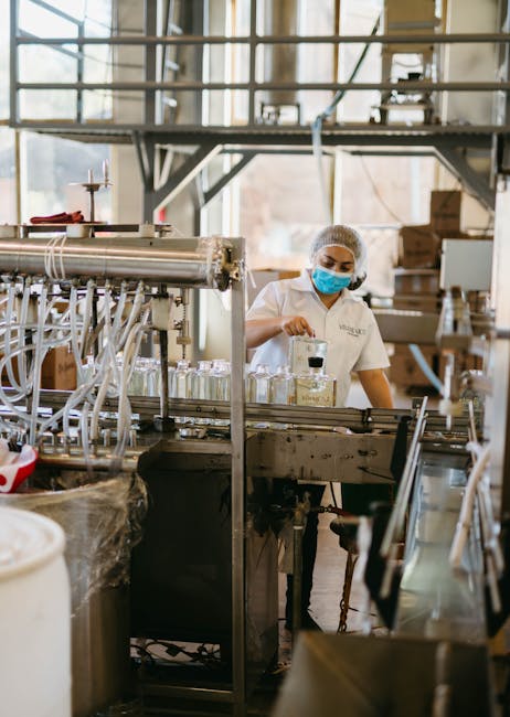 A worker wearing a mask and hairnet operates machinery in a tequila production facility.